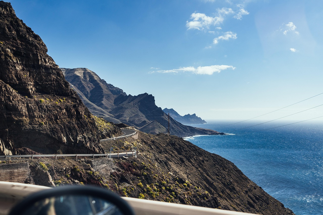 Driving along a winding coastal road on Gran Canaria, with panoramic views of cliffs, ocean, and sunlit landscape